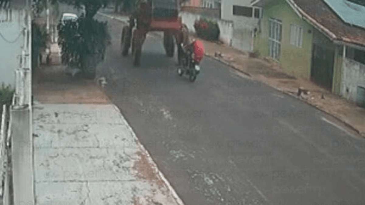 Imagem colorida mostra uma moto, com duas pessoas, em uma rua, e uma máquina agrícola vindo no sentido oposto antes de um acidente acontecer.